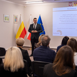 Na sali konferencyjnej zastępca prezydenta Tomasz Klim przemawia do zgromadzonej publiczności. W tle widoczne flagi Polski i Unii Europejskiej oraz prezentacja na ekranie.