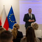 Zastępca prezydenta Tomasz Klim przemawia do grupy osób siedzących na sali, w tle widoczne flagi Polski i Unii Europejskiej oraz prezentacja na ekranie.
