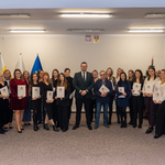 Grupa osób stoi w sali, trzymając dyplomy. W tle flagi i choinka. Wszyscy uśmiechają się do kamery.