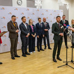 Mężczyzna stoi przed mikrofonami podczas konferencji prasowej, w tle stoją inni uczestnicy.