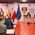 Kobieta stoi przy mównicy, radni siedzą przy biurkach.