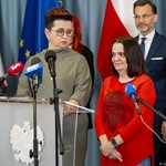 Kobieta stoi przy mównicy podczas konferencji prasowej, w tle stoją inni uczestnicy.