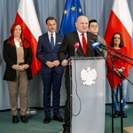 Mężczyzna stoi przy mównicy, w tle stoją inni uczestnicy.