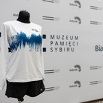 Biały t-shirt z niebieskim nadrukiem, w tle: ścianka z logiem Miasta Białegostoku oraz Muzeum Pamięci Sybiru 