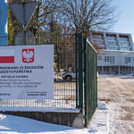 Baner z informacją o dofinansowaniu ze środków budżetu państwa wisi na ogrodzeniu Miejskiego Ośrodka Sportowego, w tle: budynek basenu oraz parking, na którym stoją zaparkowane auta