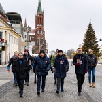 Prezydent Tadeusz Truskolaski, Poseł na Sejm Krzysztof Truskolaski, Zastępca prezydenta Rafał Rudnicki oraz inni wolontariusze idą chodnikiem Rynku Kościuszki trzymając w dłoniach puszki WOŚP. W tle: choinka 