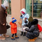 Mały chłopiec ubrany w pomarańczową kurtkę i zimową czapkę wkłada pieniądze do puszki WOŚP trzymanej przez dwie wolontariuszki 