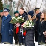Mundurowi oraz uczestnicy wydarzenia stoją przed pomnikiem trzymając w rękach wieńce zrobione z białych kwiatów