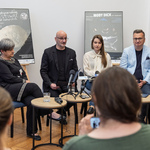 Uczestnicy konferencji prasowej siedzą przy stoliczkach na których stoją mikrofony.
