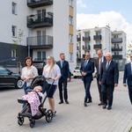 Uczestnicy konferencji prasowej idą spacerem chodnikiem, w tle zabudowania.
