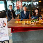 Uczestnicy konferencji prasowej siedzą przy blacie w restauracji, obok stoi plakat promujący wydarzenie.