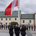 Trzech funkcjonariuszy Straży Miejskiej stoi przy maszcie na którym zawieszono flagę Polski.