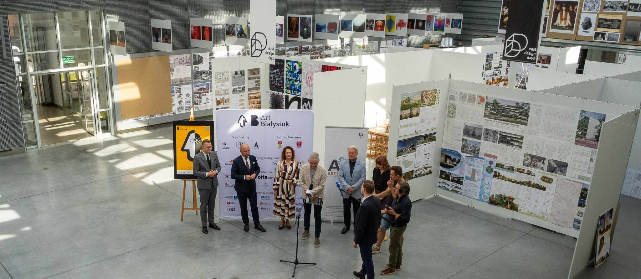 Uczestnicy konferencji stoją w dużej sali, w tle wystawa zdjęć i obrazów.