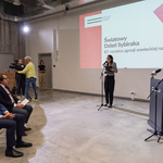 Uczestnicy konferencji siedzą na krzesłach, przed nimi z mikrofonem stoi prowadząca wydarzenie.