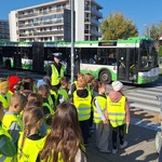 Uczniowie w kamizelkach odblaskowych stoją przed przejściem dla pieszych. W tle policjantka oraz autobus komunikacji miejskiej