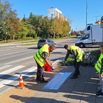 Dzieci w kamizelkach odblaskowych stoją przed przejściem dla pieszych, służba drogowa maluje napis na chodniku 