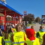 Uczniowie w kamizelkach odblaskowych wręczają na przystanku opaski odblaskowe, osobom czekającym na autobus