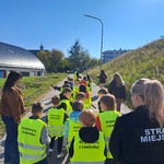 Uczniowie w kamizelkach odblaskowych idą gęsiego w dwurzędzie, obok opiekunowie