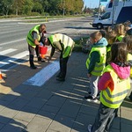 Dzieci w kamizelkach odblaskowych stoją przed przejściem dla pieszych, służba drogowa maluje napis na chodniku 