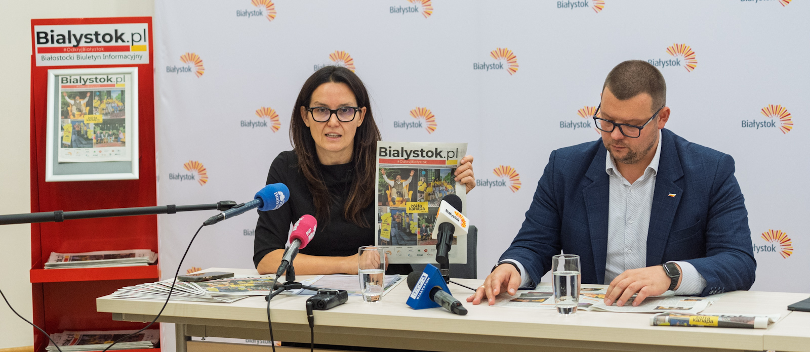 Przedstawiciele Miasta siedzą przed mikrofonami podczas konferencji prasowej.