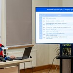 Dyrektorka CAS Urszula Przystupa mówi do mikrofonów podczas konferencji prasowej, w tle stoi monitor z wyświetlaną grafiką.
